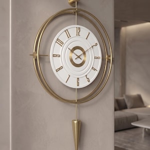 Può includere: Un orologio da parete con quadrante circolare bianco, numeri dorati e un centro dorato testurizzato. L'orologio è racchiuso da due anelli metallici concentrici dorati e presenta un pendolo conico dorato che pende verso il basso.