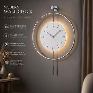 Può includere: Un orologio da parete circolare con quadrante bianco, lancette argentate e numeri nelle posizioni 3, 6 e 12. L'orologio è sospeso all'interno di una sottile cornice metallica circolare e presenta un'asta verticale con un peso cilindrico.