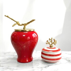 May include: Two decorative ceramic jars. One is a glossy red urn with a gold branch-shaped lid handle. The other is a small, round jar with red and white stripes and a gold coral-shaped lid handle. Both are on a marble surface.