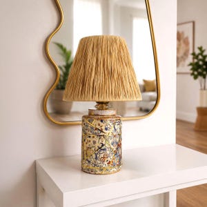 May include: A table lamp featuring a cylindrical ceramic base with a multi-coloured mosaic pattern of blue, red, and gold accents, accented by gold lines. The lamp is topped with a conical, textured, light brown raffia lampshade.