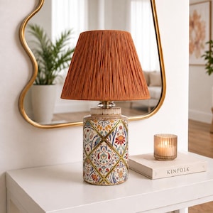 May include: A table lamp with a pleated, burnt-orange fabric shade and a cylindrical ceramic base featuring a colourful, repeating diamond-shaped pattern of floral and geometric motifs in shades of blue, red, yellow, and green on a white background.