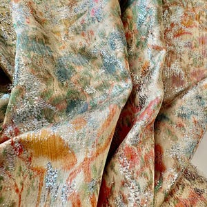 May include: Close-up of a fabric with a metallic sheen. The fabric has a floral pattern in shades of orange, pink, green, and blue. The fabric is textured and has a subtle shimmer.