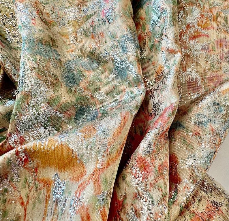 May include: Close-up of a fabric with a metallic sheen. The fabric has a floral pattern in shades of orange, pink, green, and blue. The fabric is textured and has a subtle shimmer.