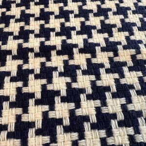 May include: Close-up of a woven fabric with a classic houndstooth pattern. The design features a repeating pattern of dark navy blue and cream-colored interlocking shapes, creating a textured surface.