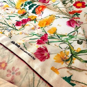 May include: White fabric with a floral print featuring pink, yellow, and red flowers. The fabric has a gold metallic thread woven into the design. The fabric has a label that reads "Alta Moda tessuto esclusivo Made in Italy".