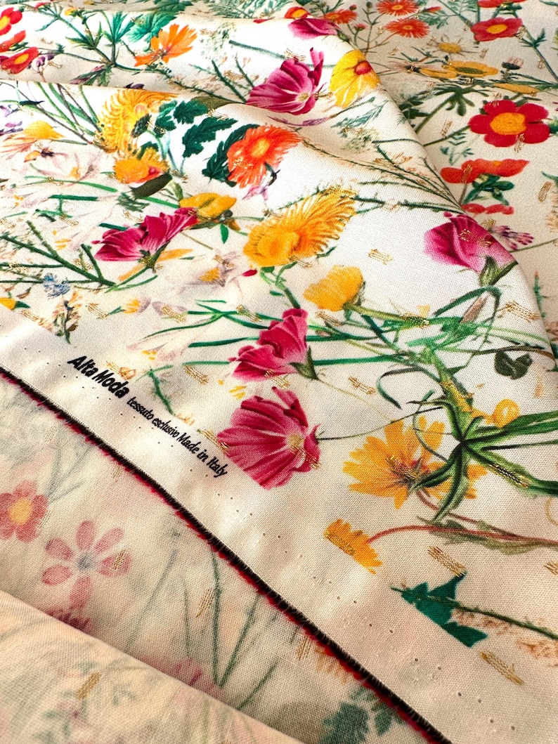 May include: White fabric with a floral print featuring pink, yellow, and red flowers. The fabric has a gold metallic thread woven into the design. The fabric has a label that reads "Alta Moda tessuto esclusivo Made in Italy".