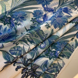 Könnte beinhalten: Weißer Stoff mit einem blauen und grünen Blumenmuster. Der Stoff zeigt eine Vielzahl von Blumen und Blättern, darunter große blaue Blumen mit weißem Zentrum und grüne Blätter mit weißem Umriss.