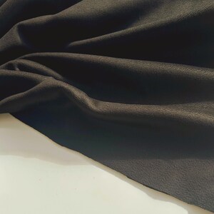 May include: A piece of black fabric with a matte, slightly textured surface. The material is draped in soft, flowing folds, creating deep shadows and highlights across the dark, opaque textile that rests against a plain, light-colored surface.