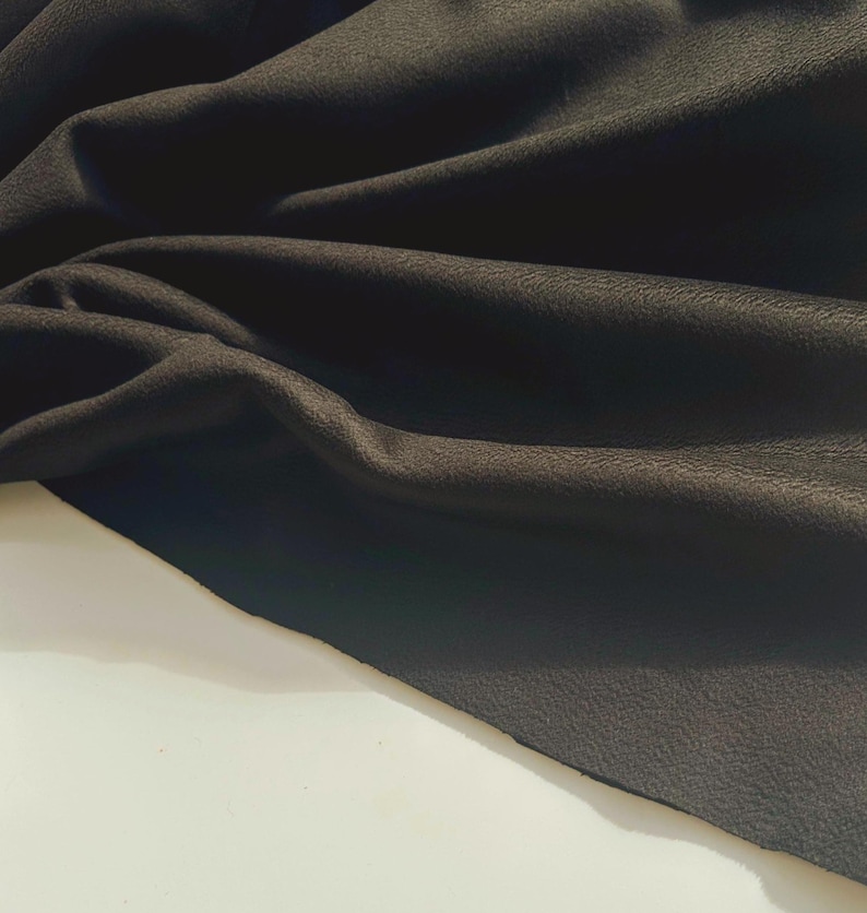 May include: A piece of black fabric with a matte, slightly textured surface. The material is draped in soft, flowing folds, creating deep shadows and highlights across the dark, opaque textile that rests against a plain, light-colored surface.