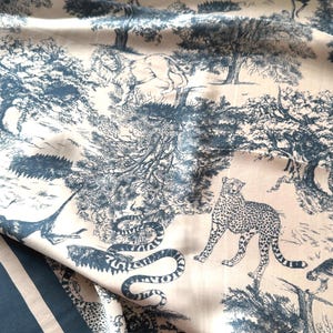 Könnte beinhalten: Cremefarbener Stoff mit blauem Toile-de-Jouy-Muster, das eine Dschungelszene mit Löwen, Leoparden und Bäumen darstellt. Der Stoff hat einen blau-weiß gestreiften Rand.