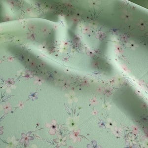 May include: A light green fabric with a delicate floral pattern. The flowers are in shades of pink, white, and blue, and they are scattered randomly across the fabric.