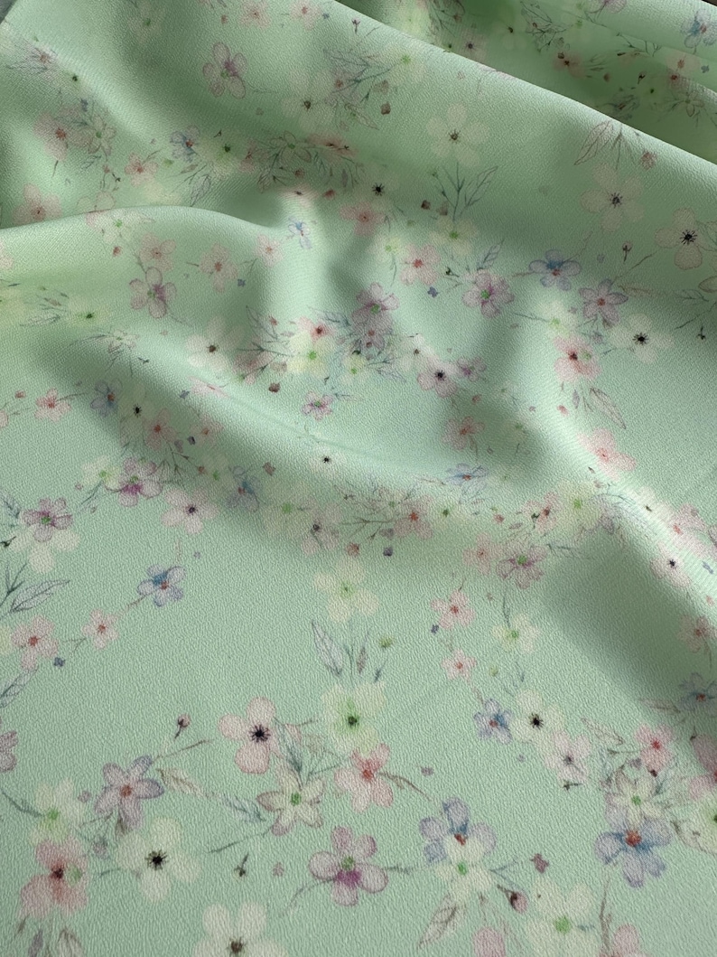 May include: A light green fabric with a delicate floral pattern. The flowers are in shades of pink, white, and blue, and they are scattered randomly across the fabric.