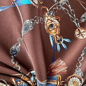 Op de afbeelding: Een bruine stof met een herhalende print van goudkleurige sieraden, waaronder ingewikkelde kettingen, sierlijke hangers met blauwe accenten en decoratieve kwastjes. Het ontwerp bevat gedetailleerde metalen schakels en ronde bedels op een bruine achtergrond.