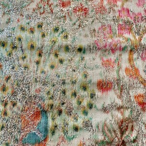 May include: Close-up of a silver fabric with a floral pattern in shades of pink, orange, and green. The fabric has a textured, metallic sheen.