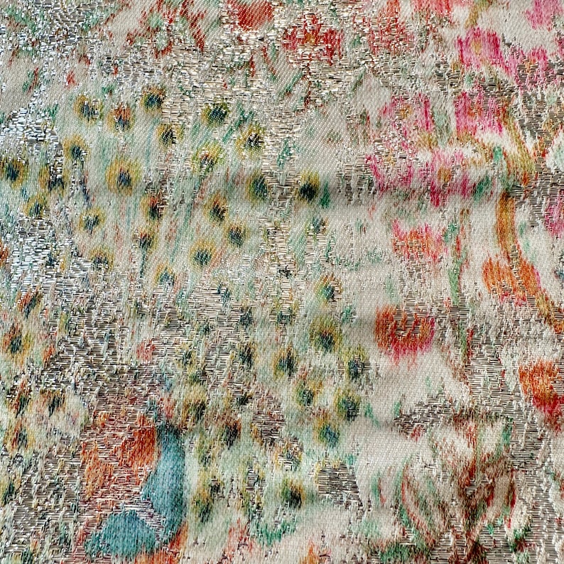 May include: Close-up of a silver fabric with a floral pattern in shades of pink, orange, and green. The fabric has a textured, metallic sheen.