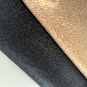 May include: Two pieces of fabric are displayed side by side. One piece is a deep black color with a textured, pebbled surface. The adjacent piece is a light tan or beige color, also featuring a consistent, fine-grained, textured surface finish.