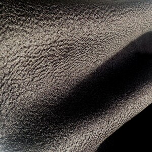 May include: A close-up view of a dark charcoal gray fabric featuring a distinct, pebbled, and highly textured surface. The material displays deep folds and shadows, highlighting the uneven, granular pattern across the entire visible textile area.