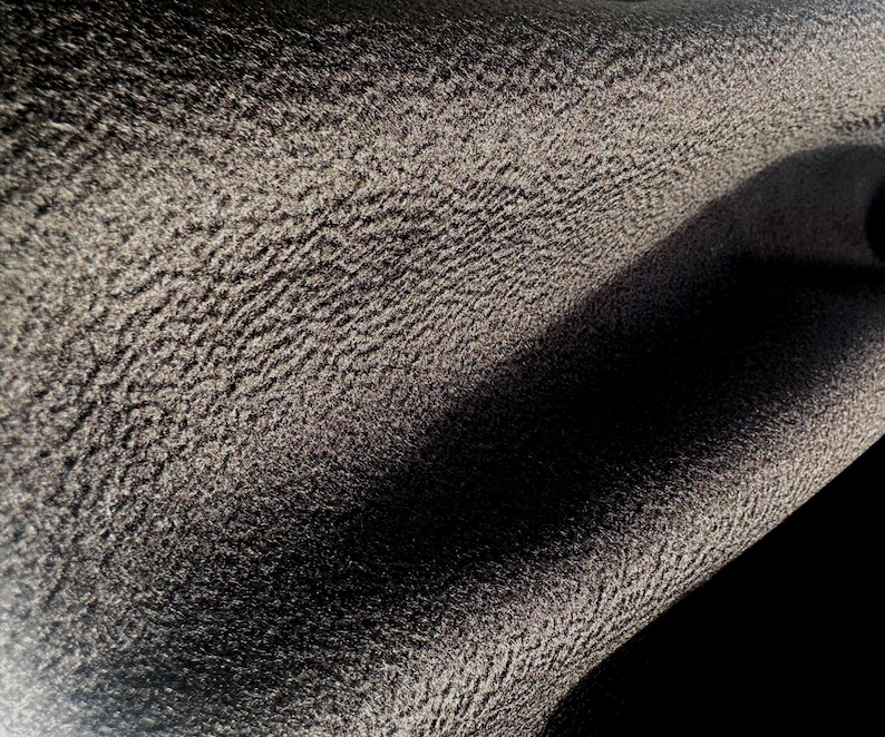 May include: A close-up view of a dark charcoal gray fabric featuring a distinct, pebbled, and highly textured surface. The material displays deep folds and shadows, highlighting the uneven, granular pattern across the entire visible textile area.