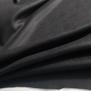 May include: A piece of black fabric with a textured, matte surface. The material is draped in soft, flowing folds, creating deep shadows and highlights across the surface. The texture appears slightly grainy and uniform throughout the entire piece.
