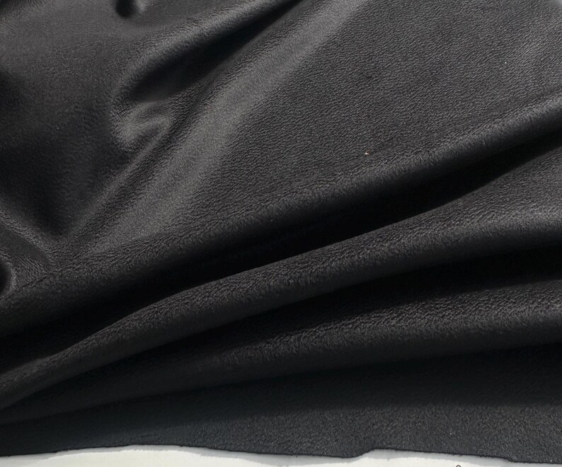 May include: A piece of black fabric with a textured, matte surface. The material is draped in soft, flowing folds, creating deep shadows and highlights across the surface. The texture appears slightly grainy and uniform throughout the entire piece.