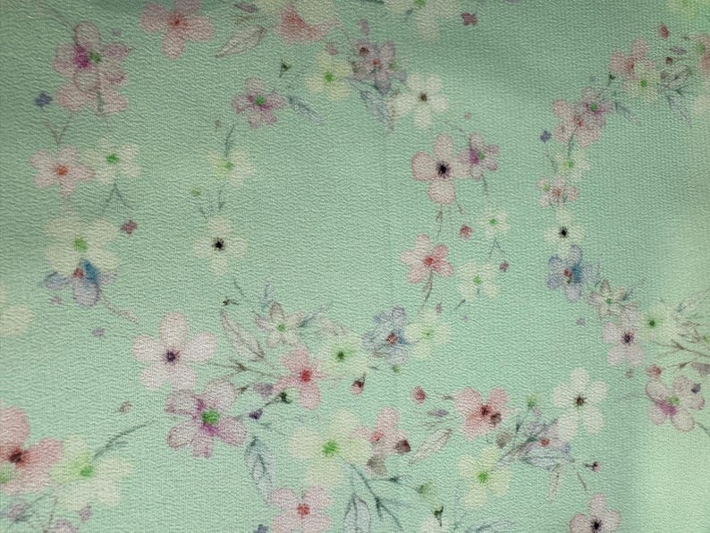 May include: A light green fabric with a delicate floral pattern. The flowers are pink, white, and blue, and they are scattered randomly across the fabric.