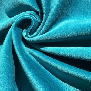 Op de afbeelding: Close-up van een zachte, turquoise blauwe fluwelen stof. De stof is gevouwen en gedrapeerd, waardoor de textuur en het luxueuze gevoel zichtbaar worden.