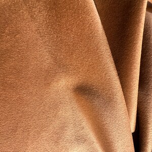 May include: A piece of fabric with a soft, matte, and suede-like texture in a warm, medium-brown color. The material is draped in folds, creating subtle shadows and highlights across the surface that emphasize the uniform, fuzzy nap of the textile.