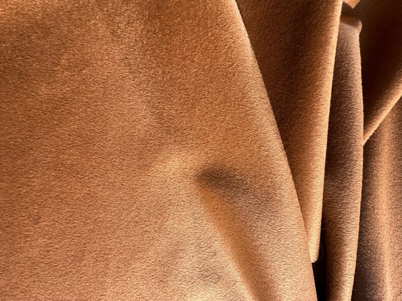 May include: A piece of fabric with a soft, matte, and suede-like texture in a warm, medium-brown color. The material is draped in folds, creating subtle shadows and highlights across the surface that emphasize the uniform, fuzzy nap of the textile.
