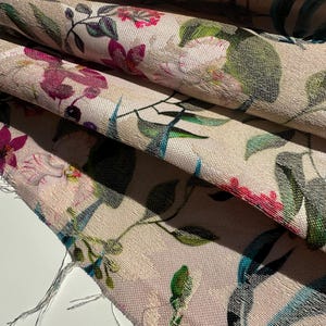 May include: Close-up of a beige fabric with a floral print. The fabric features pink, white, and green flowers with blue and green leaves. The fabric has a textured surface.