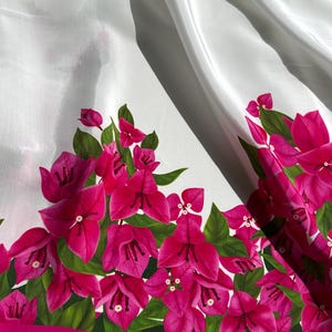 Könnte beinhalten: Weißer Stoff mit einem lebendigen Blumenrand, der leuchtend pinkfarbene Bougainvillea-Blüten und grüne Blätter zeigt. Die Blumen sind entlang eines pinkfarbenen Randes mit Fransen angeordnet und bilden einen kräftigen Kontrast zum weißen Hintergrund.
