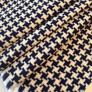 May include: Close-up of a folded fabric with a classic houndstooth pattern. The fabric features a repeating design of navy blue and off-white interlocking shapes. The edges of the fabric have a fringed detail.