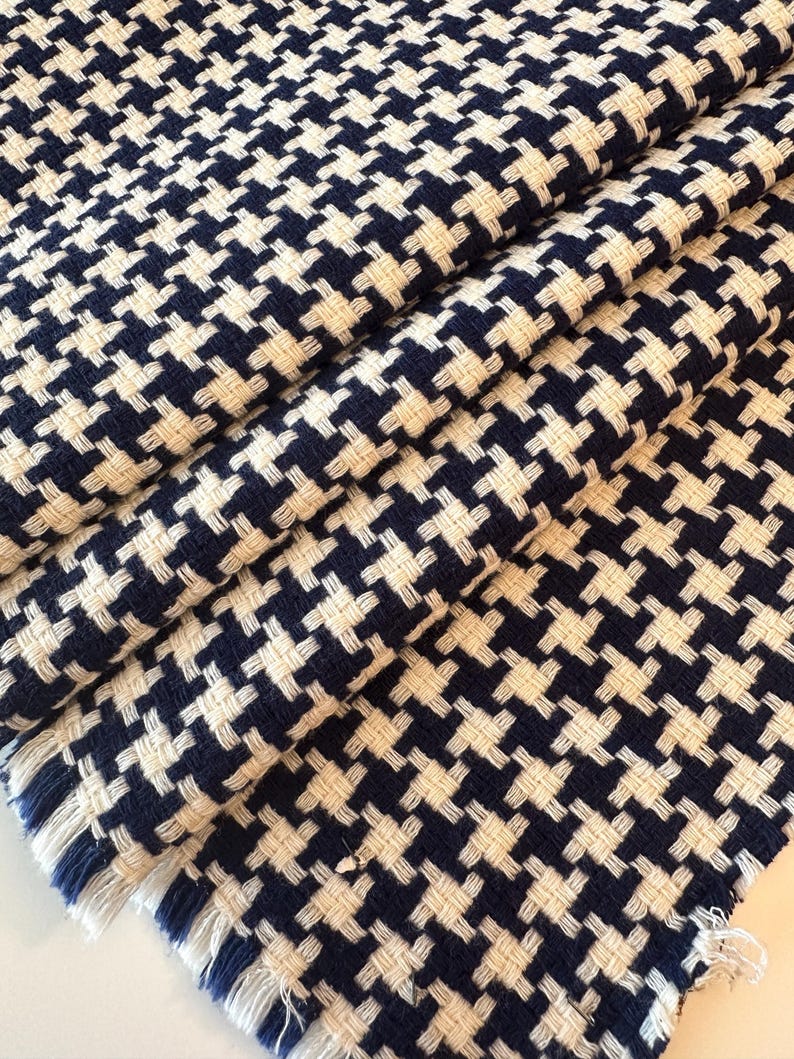 May include: Close-up of a folded fabric with a classic houndstooth pattern. The fabric features a repeating design of navy blue and off-white interlocking shapes. The edges of the fabric have a fringed detail.