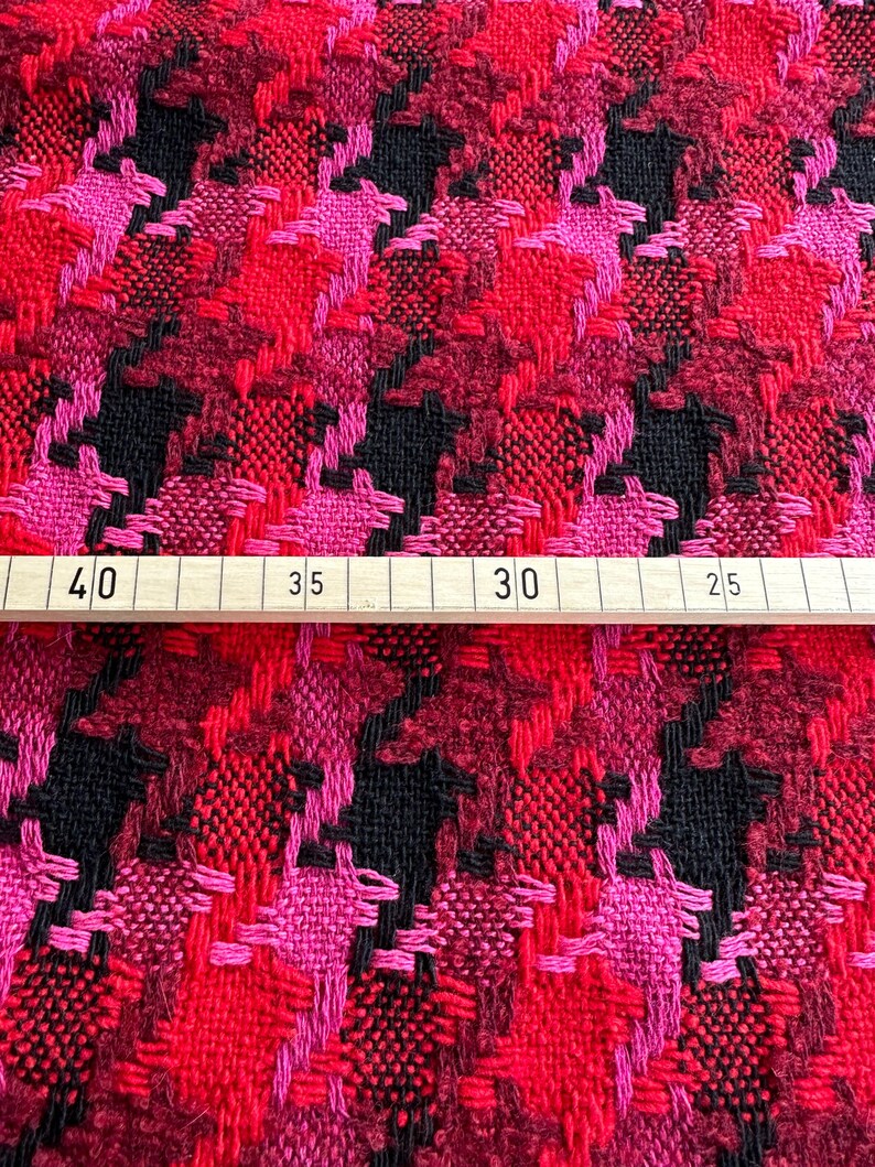 Puede incluir: Tela de pata de gallo roja, rosa y negra con textura tejida. La tela se mide con una regla que muestra 40, 35, 30 y 25.