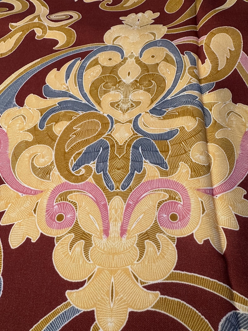 May include: A close-up of a burgundy fabric with a gold, pink, and blue damask pattern.