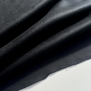 May include: A piece of black leather fabric with a visible grain texture, featuring deep folds and soft drapes that create natural shadows across the surface, showcasing the material's flexible quality and matte finish in a close-up view.
