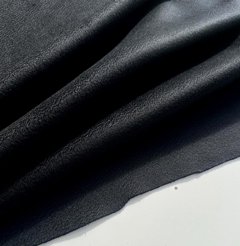 May include: A piece of black leather fabric with a visible grain texture, featuring deep folds and soft drapes that create natural shadows across the surface, showcasing the material's flexible quality and matte finish in a close-up view.