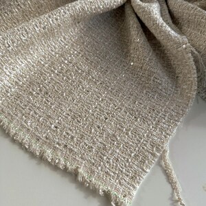 Puede incluir: Primer plano de una tela texturizada de color beige neutro, con un diseño tejido. La tela tiene un brillo sutil y está adornada con pequeñas lentejuelas dispersas. El borde de la tela está deshilachado, con un delicado ribete colgando de la esquina.
