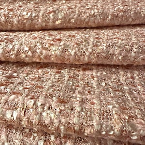 Puede incluir: Una tela texturizada que presenta una mezcla tejida de hilos de color rosa claro, crema y oro rosa metálico. El material muestra un tejido grueso y multidimensional con un patrón de cuadrícula consistente en toda la superficie.