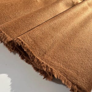 May include: Close-up of a folded, textured fabric in a warm, earthy brown hue. The fabric has a soft, furry appearance with a fringed edge, suggesting a textile like faux fur or a similar material. The folds create shadows.
