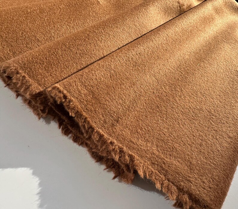 May include: Close-up of a folded, textured fabric in a warm, earthy brown hue. The fabric has a soft, furry appearance with a fringed edge, suggesting a textile like faux fur or a similar material. The folds create shadows.