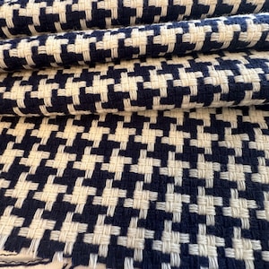 May include: Close-up of a folded fabric with a houndstooth pattern. The fabric features a repeating design of navy blue and off-white woven squares. The texture of the fabric is visible, with the threads creating a slightly raised surface. The fabric is likely used for upholstery or clothing.