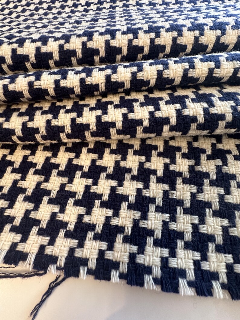 May include: Close-up of a folded fabric with a houndstooth pattern. The fabric features a repeating design of navy blue and off-white woven squares. The texture of the fabric is visible, with the threads creating a slightly raised surface. The fabric is likely used for upholstery or clothing.