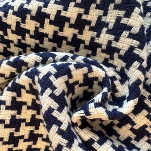 May include: Close-up of a woven fabric with a houndstooth pattern. The fabric features a repeating design of navy blue and off-white squares. The material is folded and draped, showing the texture and weave of the fabric.