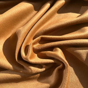 May include: Close-up of a folded, textured fabric in a warm, golden-brown hue. The material has a soft, velvety appearance with visible folds and shadows, suggesting a luxurious textile. The fabric's surface appears to have a slight sheen.