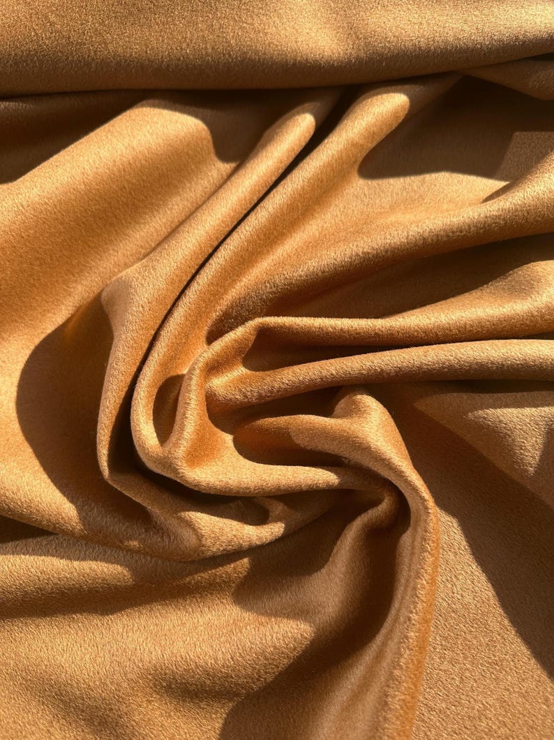 May include: Close-up of a folded, textured fabric in a warm, golden-brown hue. The material has a soft, velvety appearance with visible folds and shadows, suggesting a luxurious textile. The fabric's surface appears to have a slight sheen.