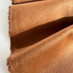 May include: A length of solid, medium-brown fabric with a soft, fuzzy, and dense texture. The material is folded in layers, revealing a raw, frayed edge along the side. The surface has a consistent, matte appearance with visible nap.