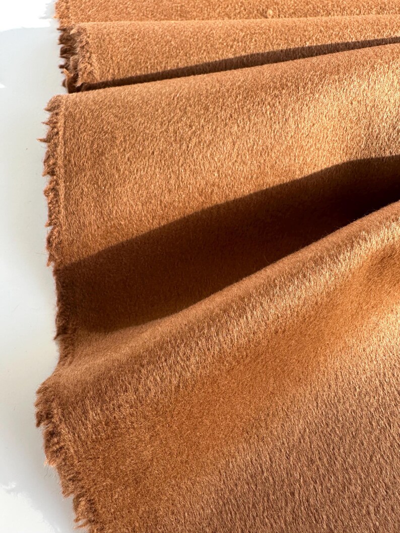May include: A length of solid, medium-brown fabric with a soft, fuzzy, and dense texture. The material is folded in layers, revealing a raw, frayed edge along the side. The surface has a consistent, matte appearance with visible nap.