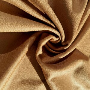 May include: A piece of tan fabric with a textured surface, arranged in a swirling, gathered pattern. The material features a soft, matte finish with deep folds and shadows created by the light, highlighting the intricate, pebbled texture.