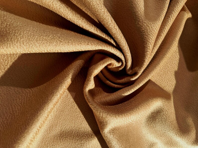 May include: A piece of tan fabric with a textured surface, arranged in a swirling, gathered pattern. The material features a soft, matte finish with deep folds and shadows created by the light, highlighting the intricate, pebbled texture.