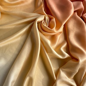 May include: A close-up of a soft, silky fabric with a gradient of colours from light yellow to orange. The fabric is draped and folded, creating a textured and flowing look.